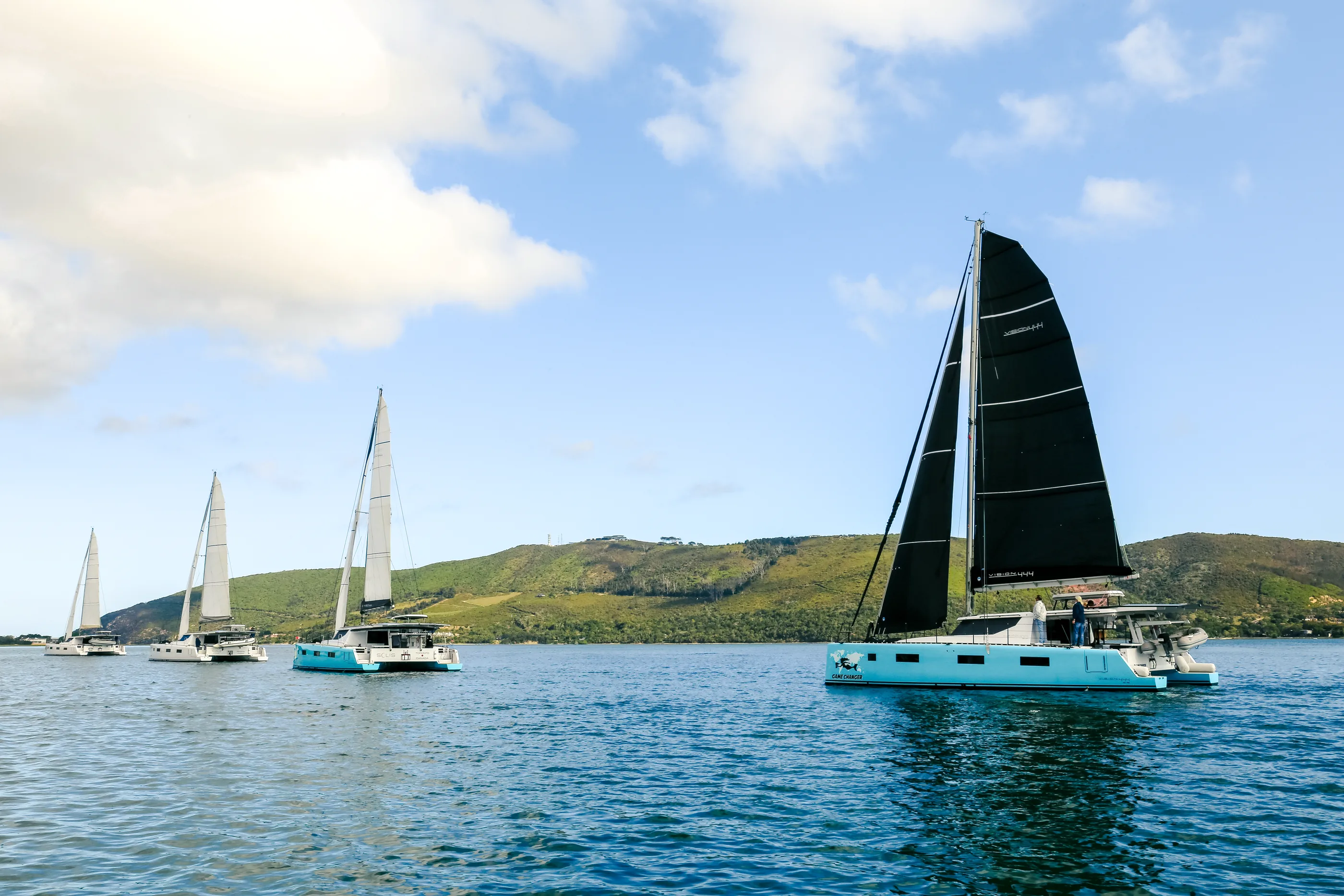 Four Vision catamarans under sail in line astern off a green coastline