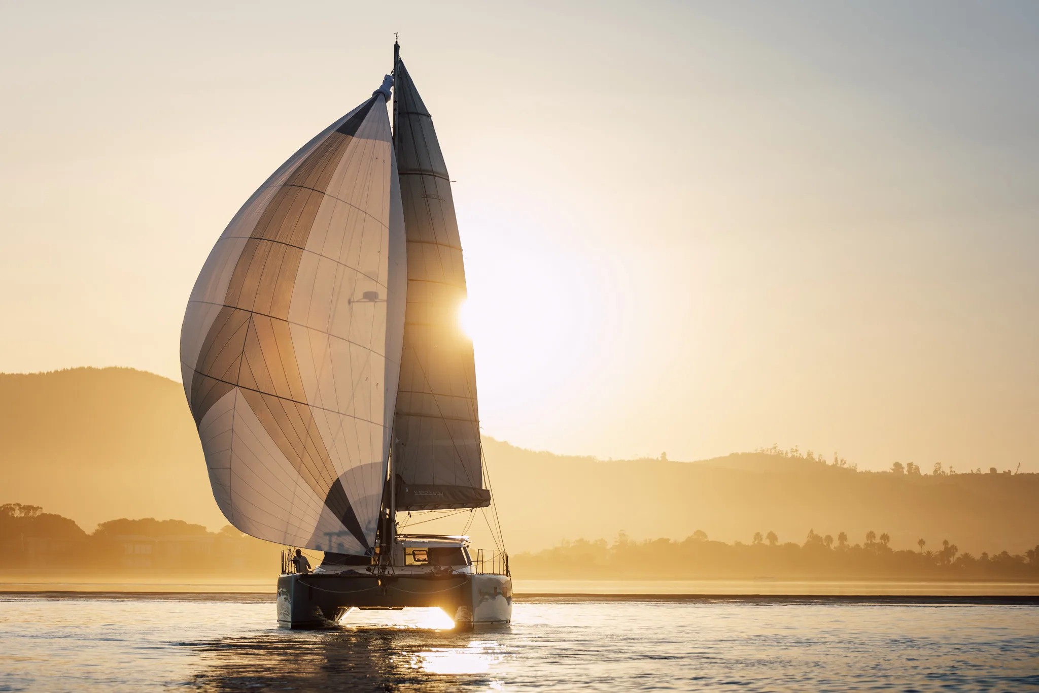 A Vision under asymmetric spinnaker at sunset, hills behind